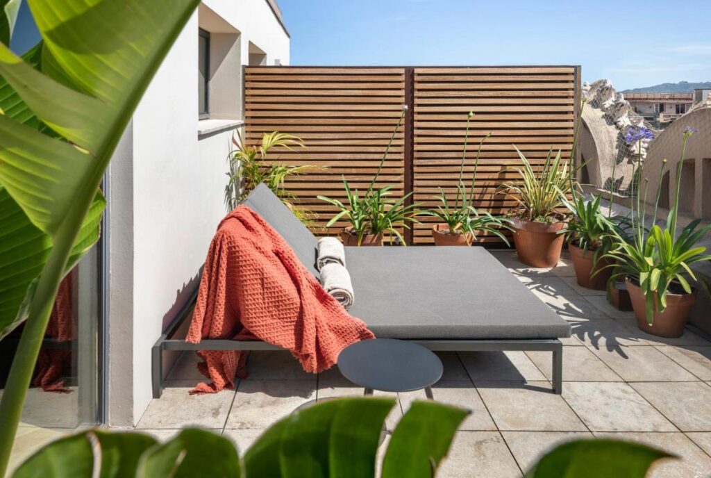 13 Unique Hotels in Barcelona, Spain 8 A modern outdoor patio at one of the unique hotels in Barcelona features a gray lounge chair, an orange throw blanket, potted plants, a sleek black side table, and a wooden privacy screen under a clear blue sky.