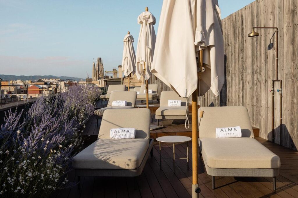 13 Unique Hotels in Barcelona, Spain 4 A rooftop terrace at one of the unique hotels in Barcelona, featuring beige lounge chairs, white Alma Hotel towels, closed umbrellas, and lavender plants. Wooden flooring complements the cityscape views under a clear sky.