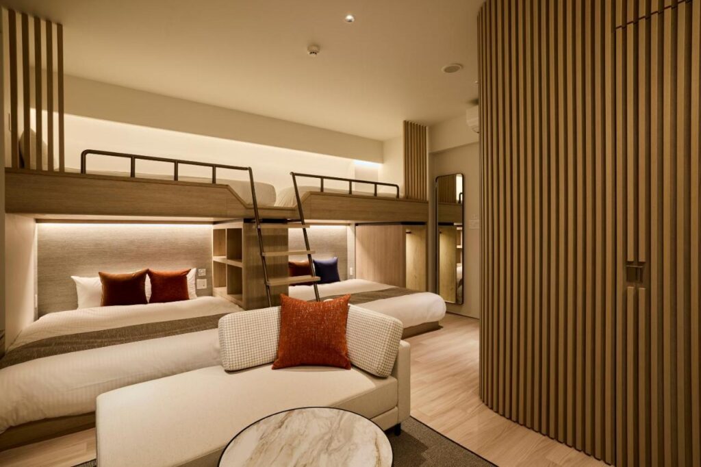 7 Best Hotels in Hiroshima, Japan 6 A modern hotel room in one of the best hotels in Hiroshima, Japan, featuring two twin beds with wooden bunk beds above, neutral decor, warm lighting, and a small round marble table with a white cushioned bench in the foreground.