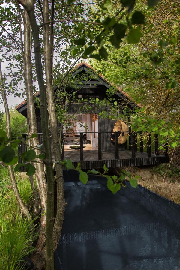 A cozy black wooden cabin with a thatched roof sits above a narrow stream, surrounded by lush green trees—like the charming escapes found in hotels in Cantabria—featuring a hanging chair on its porch.