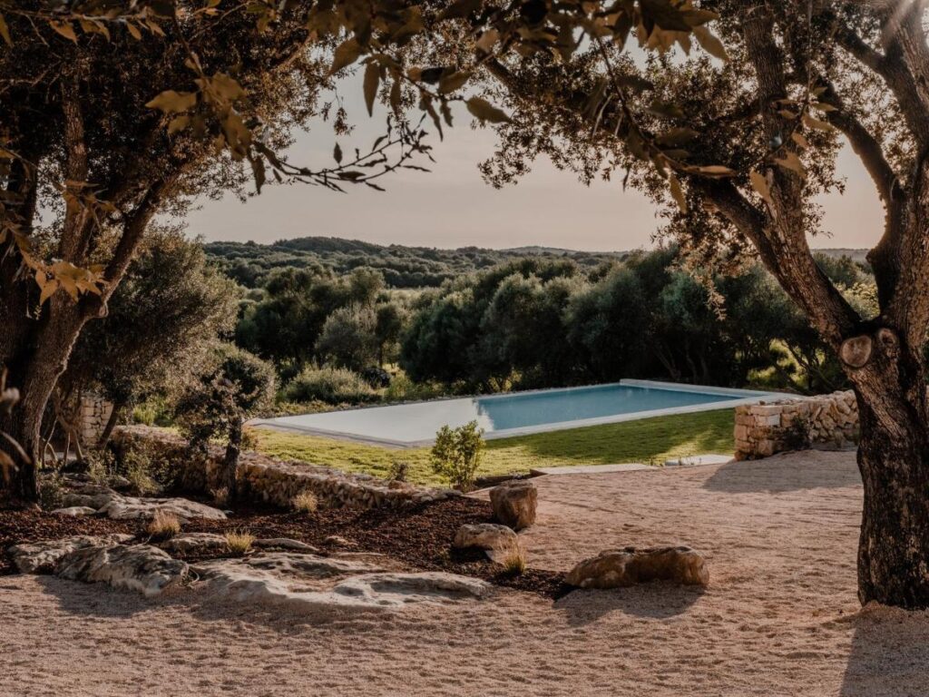 Unique Boutique Hotels in Menorca 20 A tranquil outdoor pool is surrounded by grass and stonework, nestled among trees and natural landscaping—evoking the serene atmosphere found at boutique hotels in Menorca, with rolling green hills visible under a soft, warm light.