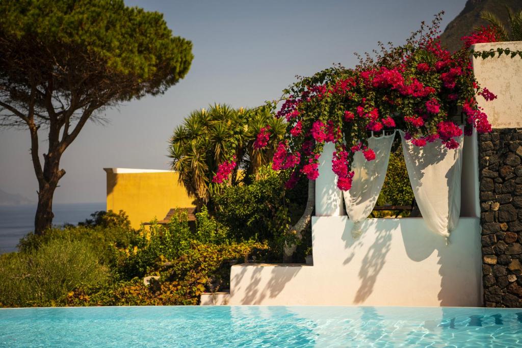 7 Best Hotels in the Aeolian Islands 29 A bright blue pool in the foreground, with white steps and draped curtains decorated with vibrant bougainvillea flowers—an inviting scene typical of hotels in the Aeolian Islands. In the background, lush greenery, a yellow building, and a glimpse of the sea under a clear sky.
