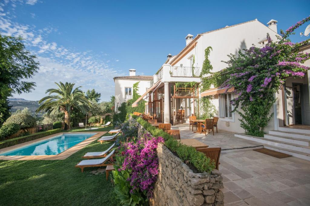 TOP 10 Luxury Hotels in Andalucía 38 Sunlit villa with ivy-covered walls, blooming purple flowers, outdoor seating, and a swimming pool surrounded by lounge chairs and lush greenery—an ambiance reminiscent of the finest luxury hotels in Andalucía under a clear blue sky.