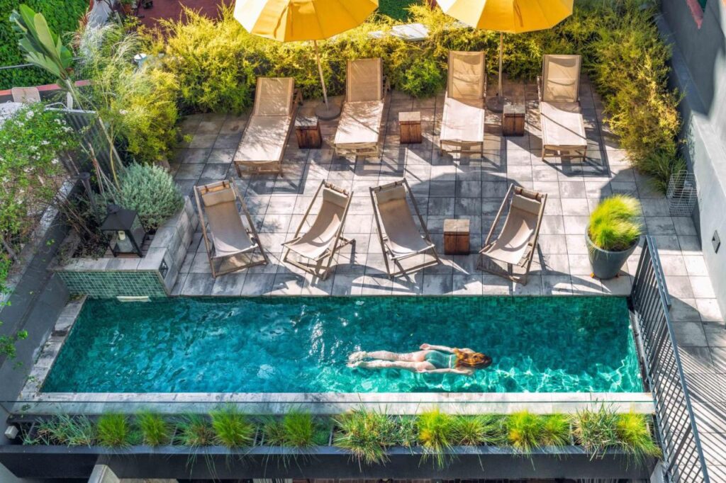 13 Unique Hotels in Barcelona, Spain 39 A woman swims alone in a narrow outdoor pool surrounded by green plants at one of the unique hotels in Barcelona. Several empty lounge chairs and two large yellow umbrellas are arranged on the sunlit tiled patio nearby.