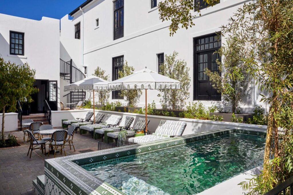 Unique Places to Stay in Cape Town 13 A stylish courtyard—a highlight among unique places to stay in Cape Town—boasts a small rectangular pool with green mosaic tiles, striped lounge chairs under white umbrellas, bistro tables, and lush greenery beside a white building with black-trimmed windows.