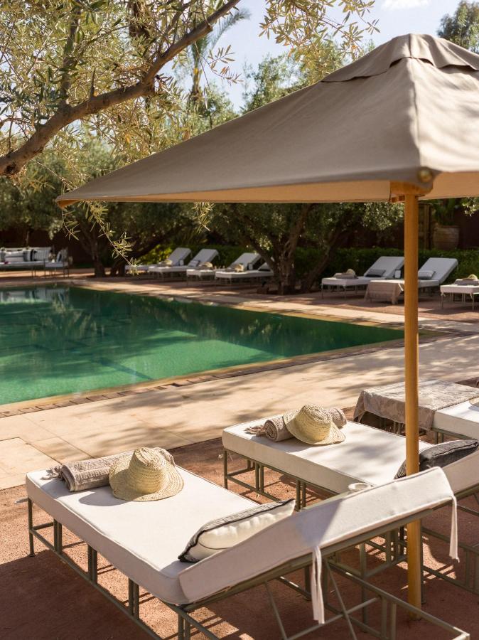 10 Unique Riads in Marrakech with Pool 22 Two poolside lounge chairs with straw hats and towels under a large beige umbrella, set by a clear swimming pool—perfect for those seeking riads in Marrakech with pool, surrounded by lush loungers and leafy trees on a sunny day.