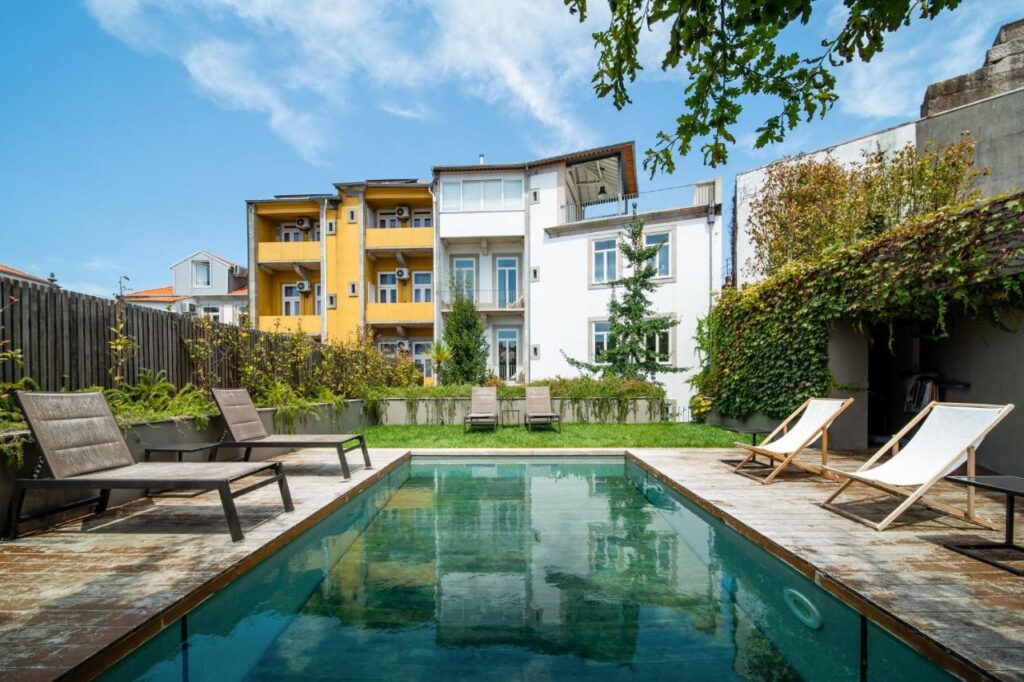 Top 12 Boutique Hotels in Porto, Portugal 37 A backyard pool with wooden lounge chairs and green grass, surrounded by lush plants—evoking the charm of boutique hotels in Porto. Modern yellow and white apartment buildings rise in the background under a clear blue sky.