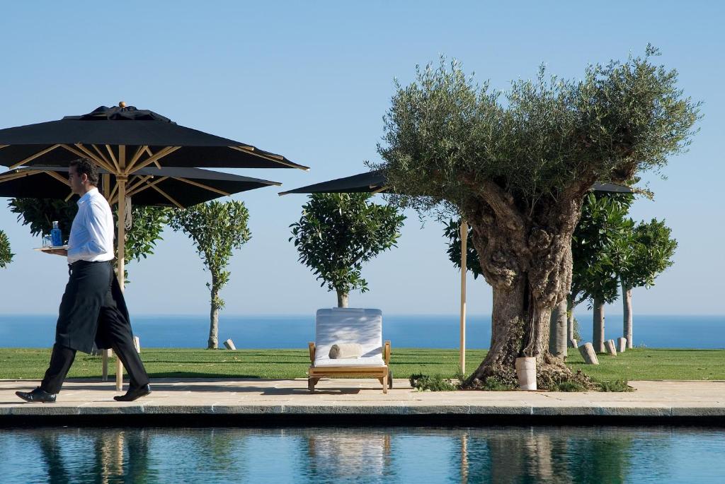 TOP 10 Luxury Hotels in Andalucía 9 A waiter carrying drinks walks by a poolside lounge chair under large black umbrellas at one of the luxury hotels in Andalucía, with green trees and the ocean visible in the background on a clear, sunny day.