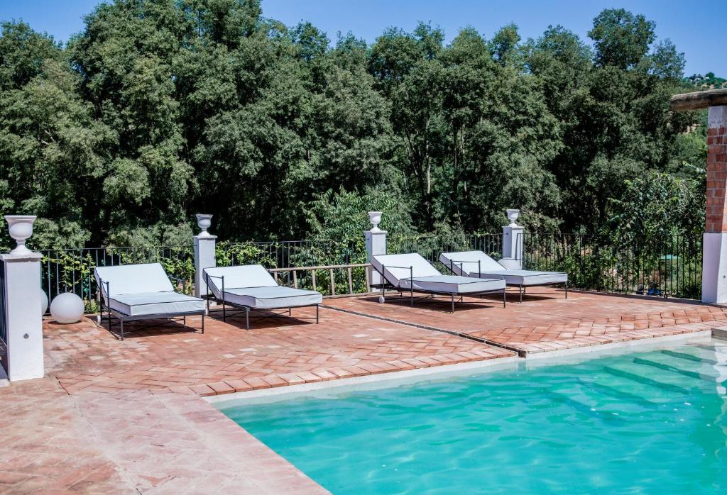 TOP 10 Luxury Hotels in Andalucía 36 Four white lounge chairs are arranged on a terracotta-tiled patio beside a swimming pool, evoking the relaxing ambiance of luxury hotels in Andalucía, with trees and greenery in the background under a clear blue sky.