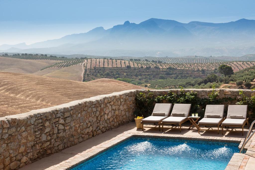 TOP 10 Luxury Hotels in Andalucía 33 A stone-walled pool with lounge chairs overlooks rolling fields, olive groves, and distant mountains under a clear blue sky—an idyllic setting found at luxury hotels in Andalucía.