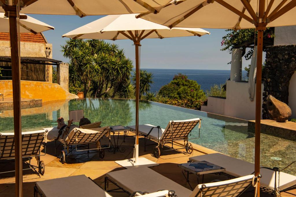7 Best Hotels in the Aeolian Islands 27 Sun loungers and umbrellas surround an infinity pool overlooking the sea at one of the charming hotels in the Aeolian Islands, with lush greenery and Mediterranean-style buildings in the background on a sunny day.