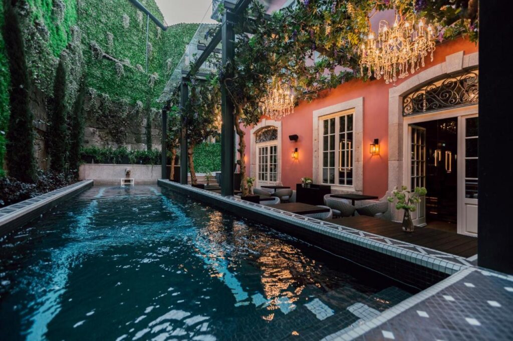 Top 12 Boutique Hotels in Porto, Portugal 8 A long, narrow pool runs alongside a pink building with large windows and ornate chandeliers hanging from a vine-covered pergola—a dreamy escape reminiscent of the boutique hotels in Porto. Lush greenery climbs the walls, with outdoor seating by the water.