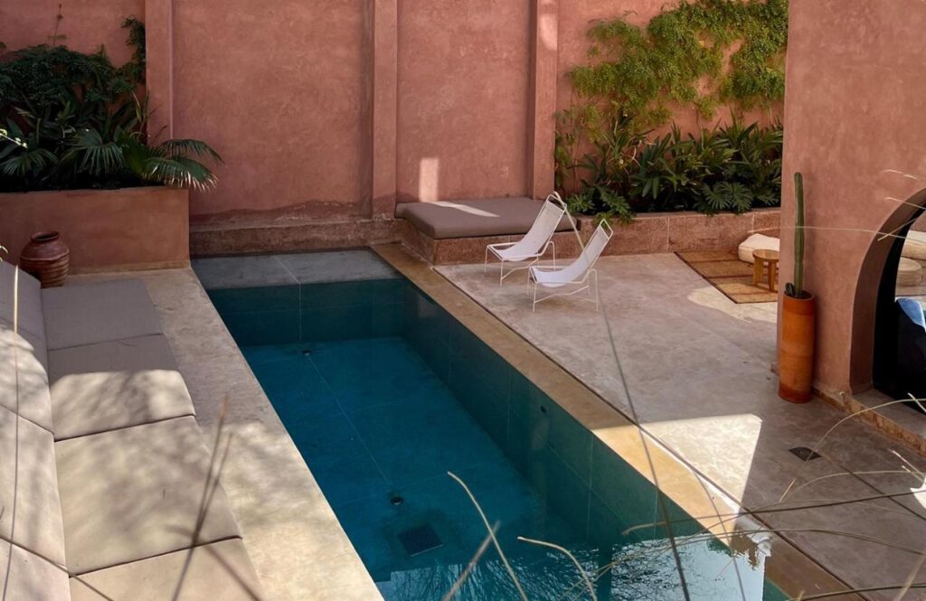 10 Unique Riads in Marrakech with Pool 8 A small rectangular pool with clear water is surrounded by beige stone, cushioned benches, two white lounge chairs, plants, and a terra cotta wall—capturing the charm of riads in Marrakech with pool in a sunlit, cozy courtyard.