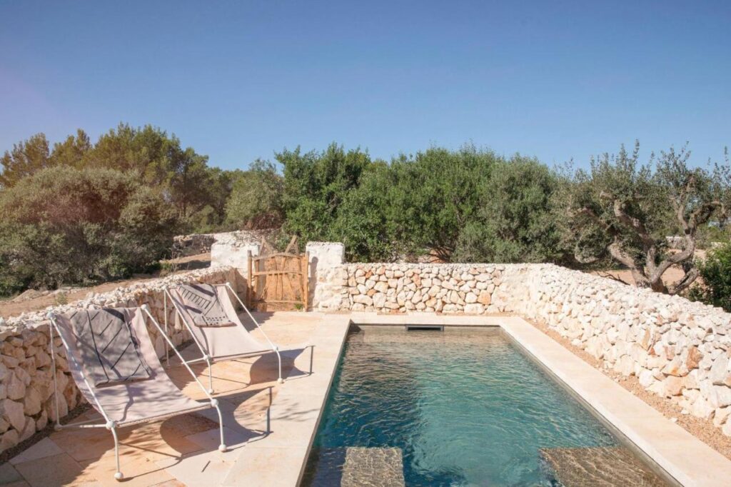 Unique Boutique Hotels in Menorca 25 A small rectangular swimming pool is surrounded by stone walls and trees, evoking the charm of boutique hotels in Menorca. Three deck chairs sit on the stone patio beside the pool, with a wooden gate visible under a clear blue sky.