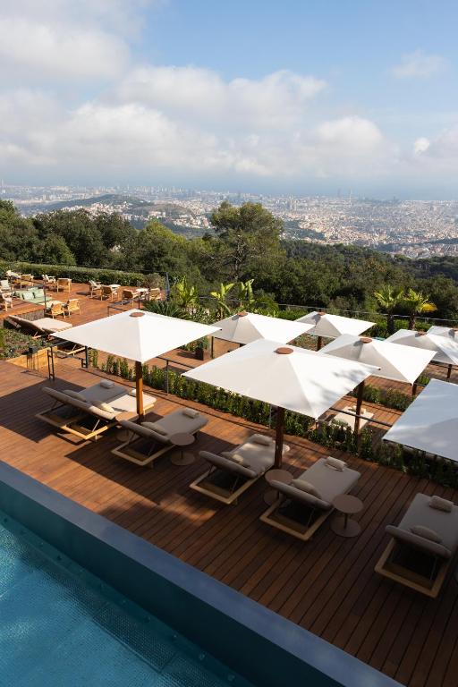13 Unique Hotels in Barcelona, Spain 46 A rooftop poolside deck at one of the unique hotels in Barcelona features lounge chairs and large white umbrellas, overlooking a cityscape surrounded by greenery and distant mountains under a partly cloudy sky.