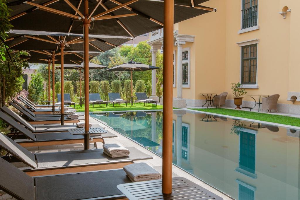 Top 12 Boutique Hotels in Porto, Portugal 50 A row of lounge chairs with towels and large umbrellas is arranged beside a narrow outdoor pool, surrounded by greenery and a yellow building—an inviting scene typical of boutique hotels in Porto.