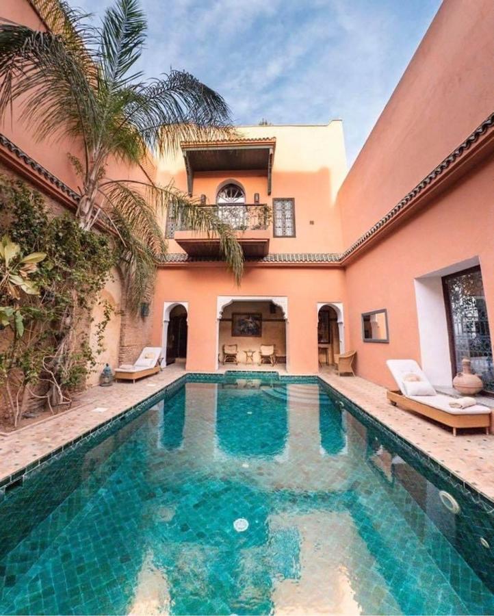 10 Unique Riads in Marrakech with Pool 3 A serene courtyard, reminiscent of riads in Marrakech with pool, features a turquoise-tiled swimming pool surrounded by peach-colored walls, palm trees, lounge chairs, and a balcony with arched windows under a blue sky.