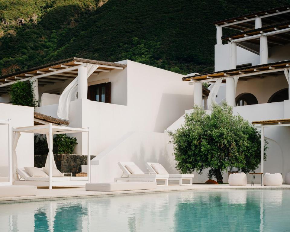 7 Best Hotels in the Aeolian Islands 6 A modern white villa with poolside lounge chairs and canopy beds, surrounded by lush greenery and mountains under a clear sky—an oasis of serenity and luxury, reminiscent of the best hotels in the Aeolian Islands.