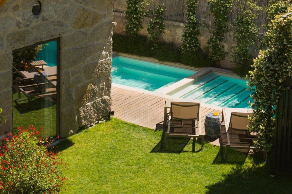 Top 12 Boutique Hotels in Porto, Portugal 25 A small backyard pool with steps sits beside a wooden deck, two lounge chairs, and green grass—reminiscent of the intimate charm found at boutique hotels in Porto. A stone building and lush plants complete this cozy outdoor setting.