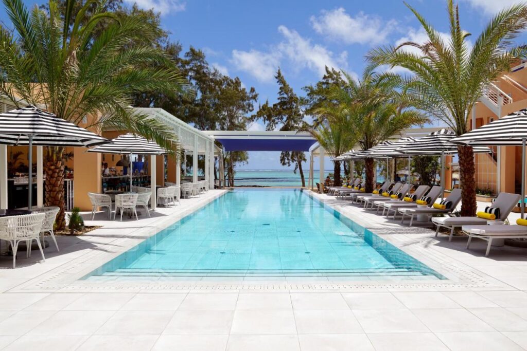 Most Unique Adults Only Hotels in Mauritius, Indian Ocean 6 A luxurious outdoor swimming pool at one of the finest adults only hotels in Mauritius, surrounded by palm trees, lounge chairs with yellow cushions, and black-and-white striped umbrellas, overlooking the ocean and blue sky.