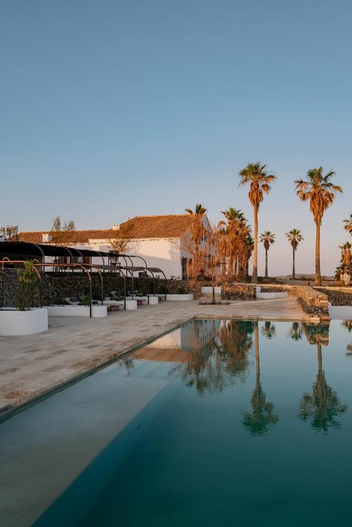 Unique Boutique Hotels in Menorca 42 A serene outdoor pool reflects palm trees and a white building with a red-tiled roof under a clear blue sky, creating the calm and inviting atmosphere found at boutique hotels in Menorca.