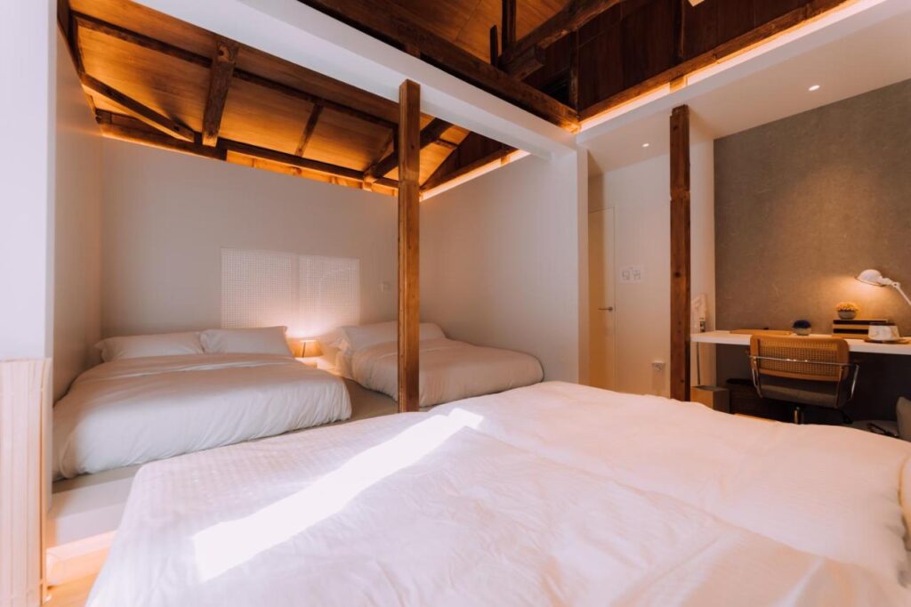15 Unique Hotels in Tokyo: Coolest hotels 54 A bright, cozy bedroom at one of the unique hotels in Tokyo, featuring two large beds, wooden beams on the ceiling, soft lighting, and a modern desk with a chair and lamp. Warm sunlight streams onto the crisp white bedding.