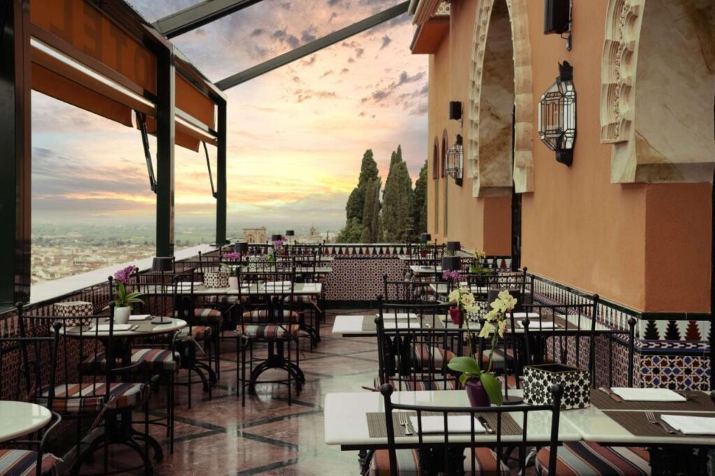 Open-air restaurant terrace with black metal tables and chairs, potted flowers on each table, patterned tiles, and arched windows. Reminiscent of boutique hotels in Granada, it overlooks a scenic city view at sunset with trees and a colorful sky.