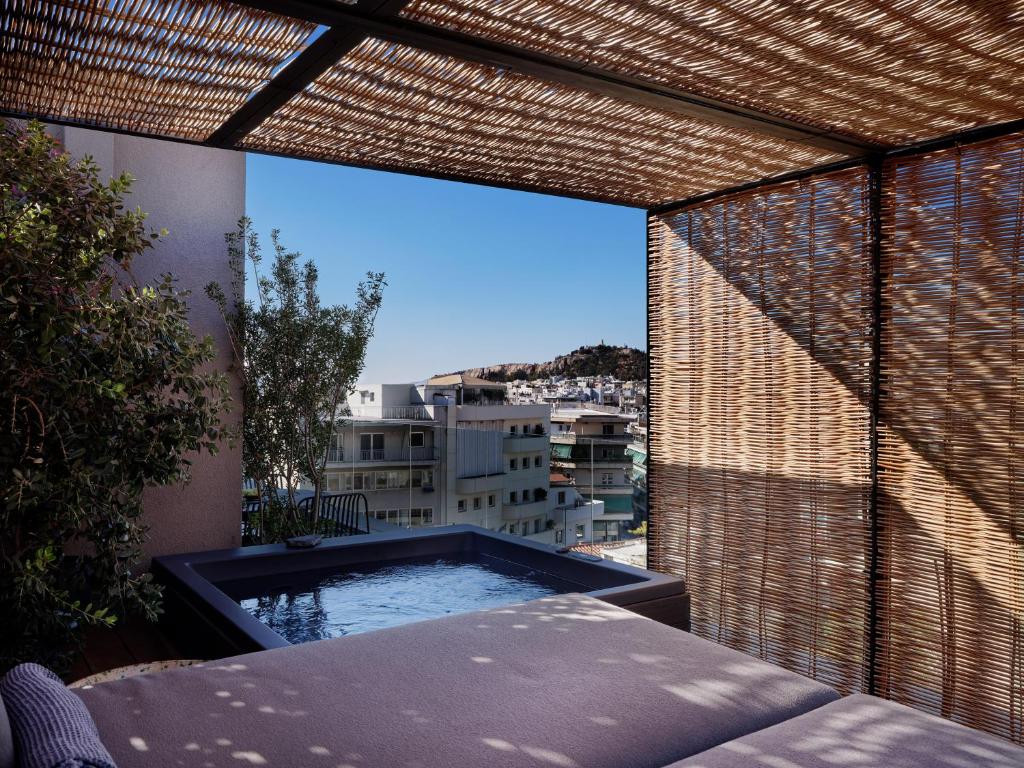A rooftop terrace at one of the boutique hotels in Athens features a small pool, lounge area, and wooden pergola with bamboo shades, offering cityscape views under a clear blue sky. Greenery and distant buildings enhance the serene setting.