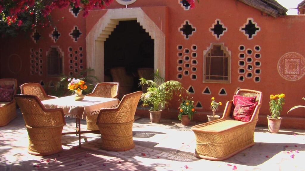 Authentic Heritage Hotels in Mandawa 7 A cozy outdoor patio at one of the charming hotels in Mandawa, featuring wicker chairs and a table set with flowers, surrounded by potted plants. The rustic terracotta wall with decorative windows adds to the inviting ambiance.
