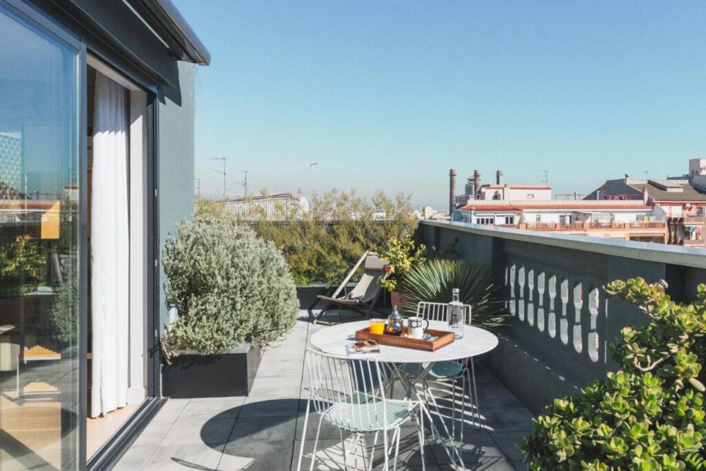 13 Unique Hotels in Barcelona, Spain 41 A sunny rooftop terrace at one of the unique hotels in Barcelona features a round table with chairs, potted plants, and lounge chairs, overlooking city buildings under a clear blue sky.