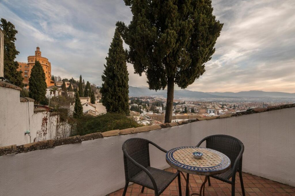 A small patio with two wicker chairs and a mosaic round table overlooks a scenic cityscape and mountains—much like the views from boutique hotels in Granada—framed by tall cypress trees and a historic orange building under a cloudy sky.