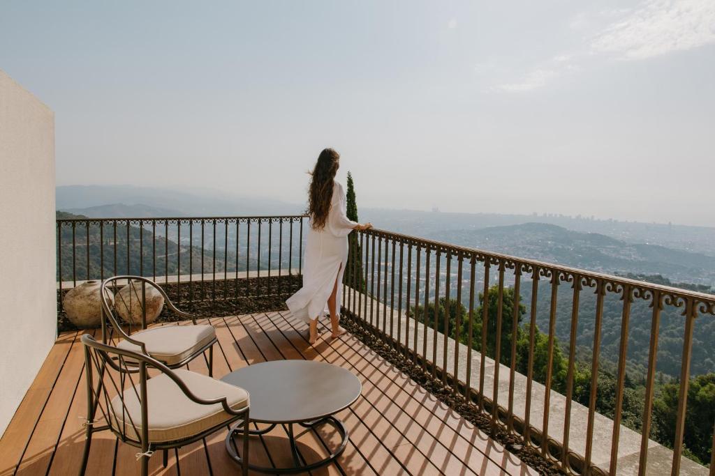 13 Unique Hotels in Barcelona, Spain 47 A woman in a white dress stands on a balcony with wooden flooring at one of the unique hotels in Barcelona, gazing over a scenic, hilly landscape and the distant city skyline beneath a hazy sky. Two chairs and a table complete the tranquil setting.