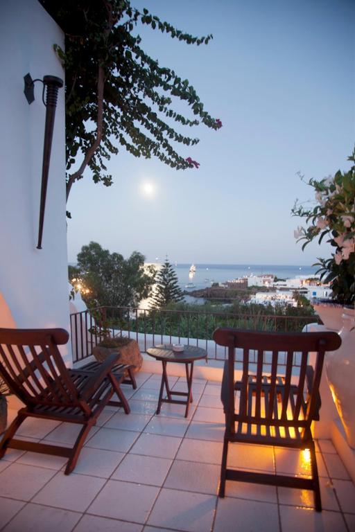 7 Best Hotels in the Aeolian Islands 25 A cozy terrace at one of the charming hotels in the Aeolian Islands features two wooden chairs and a small table, overlooking white buildings, lush greenery, and the sea at dusk, with the full moon shining above.