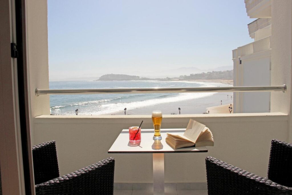 A balcony at one of the finest hotels in Cantabria overlooks the ocean, featuring a small table with a red cocktail, a glass of beer, and an open book. Two black chairs face the scenic beach and distant coastline under a clear sky.