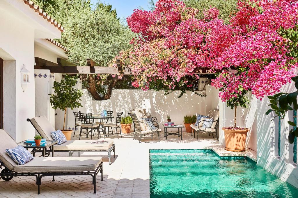 TOP 10 Luxury Hotels in Andalucía 20 A sunny patio reminiscent of luxury hotels in Andalucía features a small turquoise pool, lounge chairs with cushions, a dining table, and vibrant pink bougainvillea blooming above, all surrounded by white walls and lush greenery.