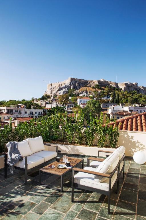A rooftop patio at one of the boutique hotels in Athens features white cushioned chairs and a coffee table, overlooking city buildings and the Acropolis under a clear blue sky.
