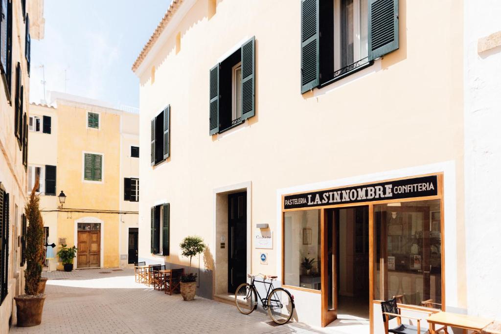 Unique Boutique Hotels in Menorca 46 Sunny street scene with yellow buildings, green shutters, and outdoor seating near Lasinombre Confiteria bakery. A bicycle rests by the entrance, potted plants line the sidewalk—just steps from charming boutique hotels in Menorca.