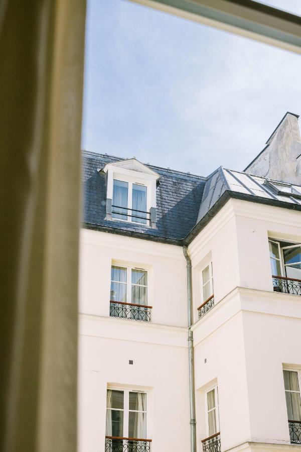 13 Unique Hotels in Paris for a Stylish Stay 24 View through a window showing the upper floors of a light-colored building with several windows, small balconies, and a slate roof under a clear blue sky—an inviting glimpse of the unique hotels in Paris.