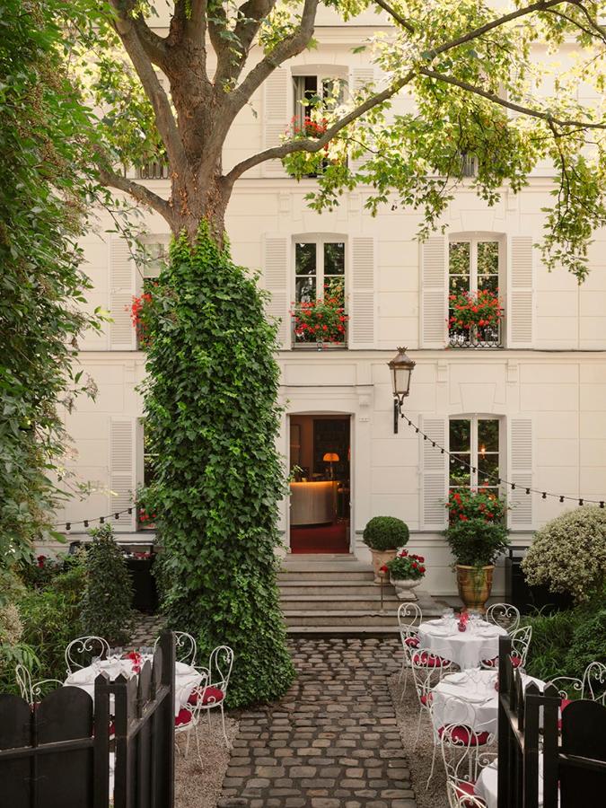 13 Unique Hotels in Paris for a Stylish Stay 9 A charming outdoor café, reminiscent of unique hotels in Paris, features white wrought iron tables and chairs with red cushions and white tablecloths, set before a white building adorned with shuttered windows, flower boxes, and greenery beneath a large tree.