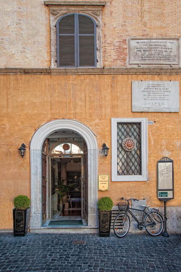 13 UNIQUE Boutique Hotels in Rome 14 An arched doorway of a historic building with yellow-orange walls, potted plants, a bicycle parked outside, wall plaques, and a menu display next to the entrance—evoking the charm of boutique hotels in Rome Italy. A shuttered window sits above.