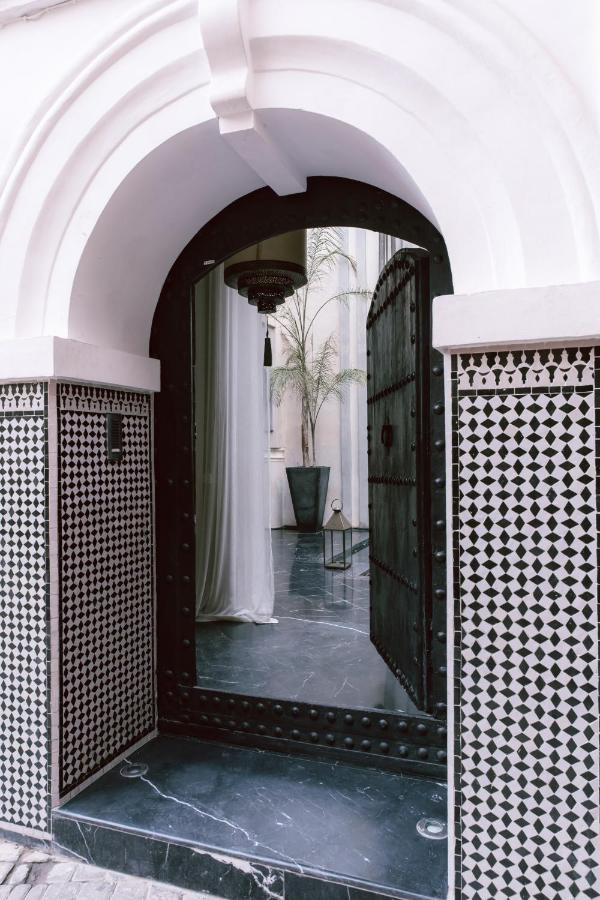 10 Unique Riads in Marrakech with Pool 28 A large arched doorway with ornate black doors opens to reveal a marble floor, a tall potted plant, a hanging chandelier, and a decorative lantern—echoing the elegance of riads in Marrakech with pool and intricate black-and-white geometric tilework.