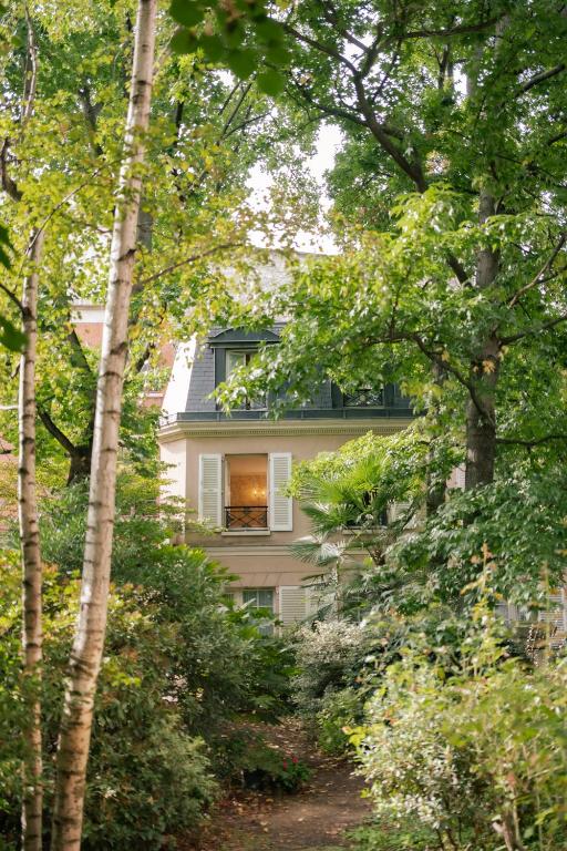 13 Unique Hotels in Paris for a Stylish Stay 42 A charming beige house with open white shutters is partially hidden behind lush green trees and dense garden foliage, exuding the peaceful, secluded ambiance often found in unique hotels in Paris.