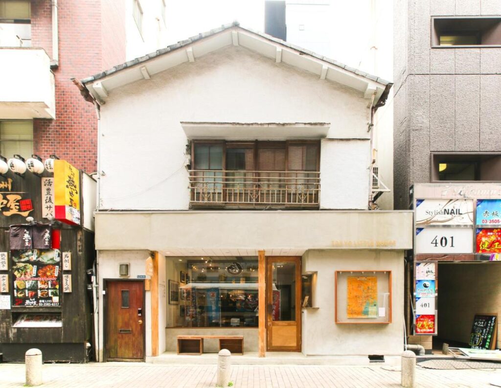 15 Unique Hotels in Tokyo: Coolest hotels 51 A two-story white building with a small balcony, flanked by taller buildings. The ground floor has large windows revealing a cozy cafe interior—neighboring storefronts feature signs and colorful ads, typical of unique hotels in Tokyo.