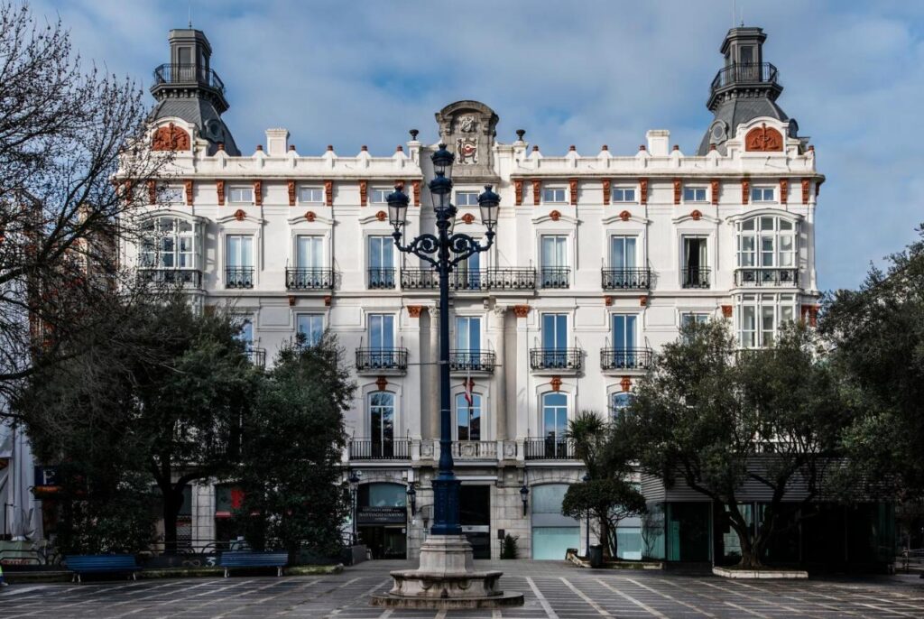 A grand, historic white building with ornate architectural details—typical of elegant hotels in Cantabria—and two towers stands behind a plaza. A decorative blue lamppost and trees frame the scene under a partly cloudy sky.