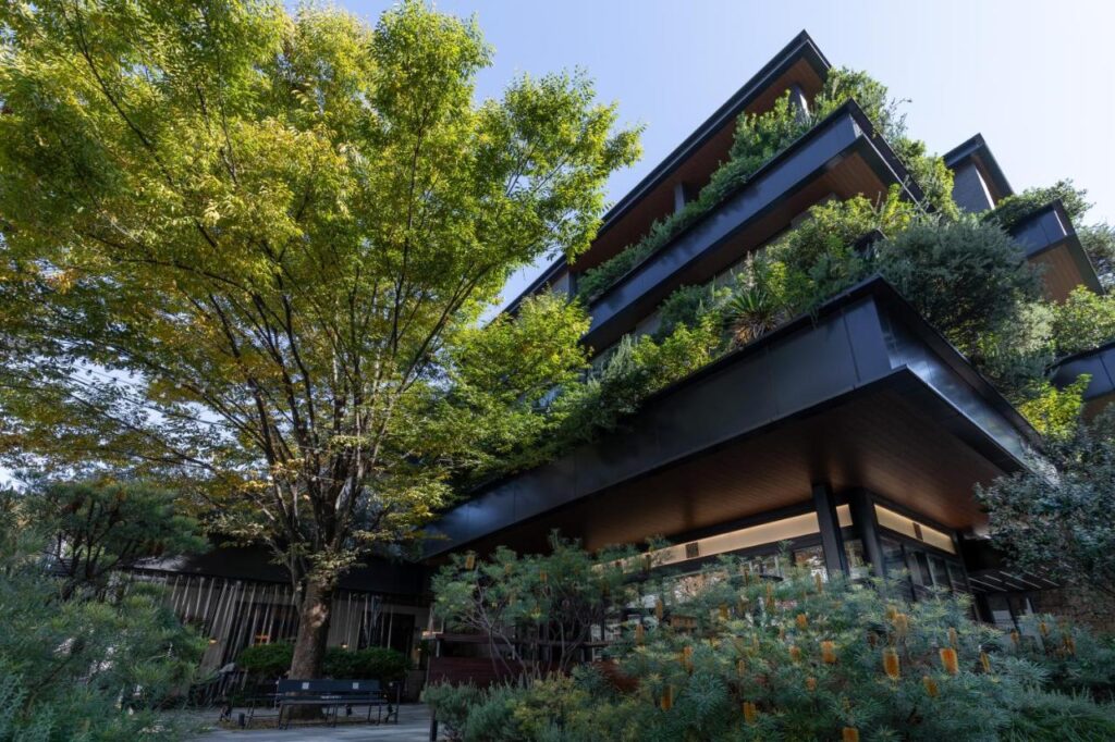15 Unique Hotels in Tokyo: Coolest hotels 6 Modern multi-story building with large horizontal balconies, each covered in lush greenery and trees—resembling one of the most unique hotels in Tokyo—nestled amid landscaped gardens and tall trees under a clear blue sky.