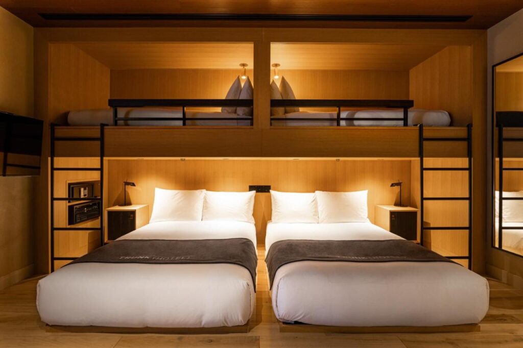 15 Unique Hotels in Tokyo: Coolest hotels 9 A modern hotel room in one of the unique hotels in Tokyo features two double beds on the lower level and two single beds in an upper loft, each accessible by ladders. The space boasts warm lighting, wood accents, and minimalistic decor.