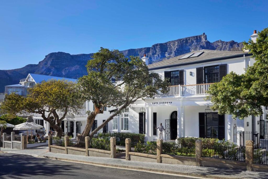 Unique Places to Stay in Cape Town 11 A white colonial-style building with black shutters and a sign reading Cape Cadogan stands on a sunny street, framed by green trees. Mountains rise in the background—one of the unique places to stay in Cape Town under a clear blue sky.
