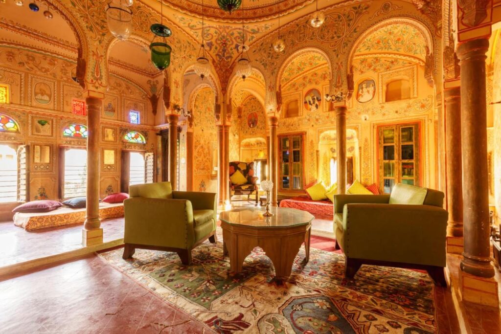 Authentic Heritage Hotels in Mandawa 4 An ornate, sunlit room with intricate arches, colorful stained glass windows, richly patterned rugs, and plush green armchairs evokes the vibrant decor of traditional Indian palace architecture found in top hotels in Mandawa.