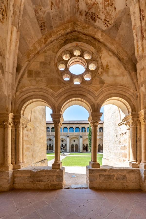 4 Best Hotels in Ribera del Duero, Spain 20 View of a medieval stone cloister with arched windows and a circular rose window, looking out onto a sunny courtyard with green grass—reminiscent of the historic charm found at some hotels in Ribera del Duero.