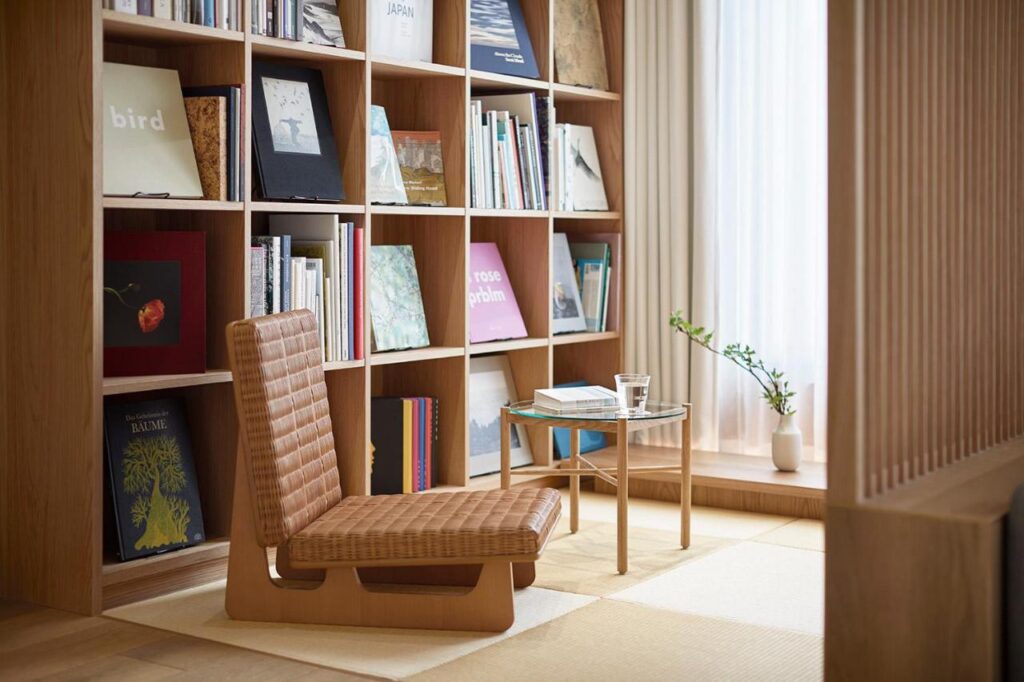 15 Unique Hotels in Tokyo: Coolest hotels 24 A cozy reading nook inspired by unique hotels in Tokyo, with a low woven seat, a small glass table holding water, and a bookshelf filled with books and art prints. Soft light filters through a sheer curtain near a potted plant.